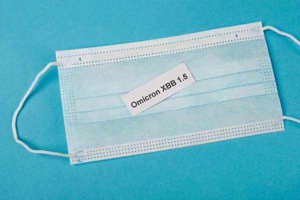 Top view of omicron xbb lettering on paper on medical mask on blue background — Stock Photo