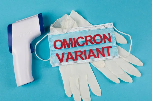 Top view of medical mask with omicron variant lettering near latex gloves and pyrometer on blue background — Stock Photo