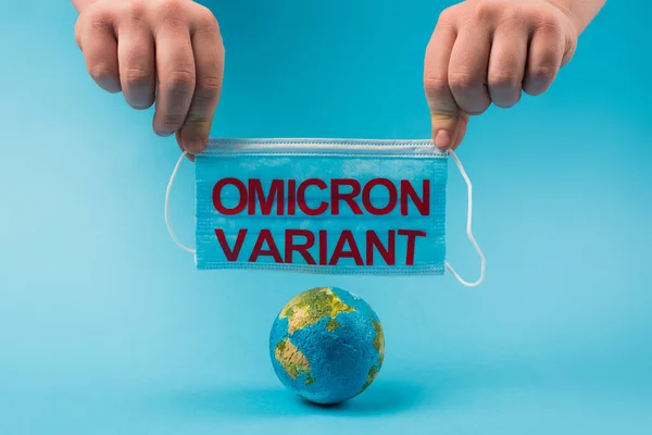 Cropped view of man holding medical mask with omicron lettering near globe on blue background — Stock Photo
