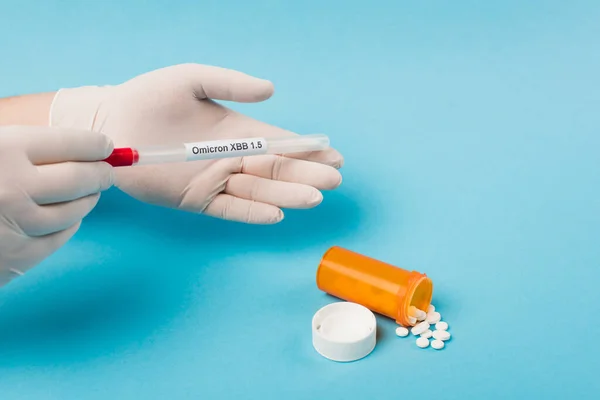 Cropped view of doctor in latex gloves holding cotton swab with omicron lettering near pills on blue background — Foto stock