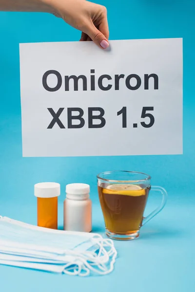 Cropped view of woman holding paper with omicron xbb lettering near blurred medical masks and cup of tea on blue background — Stock Photo