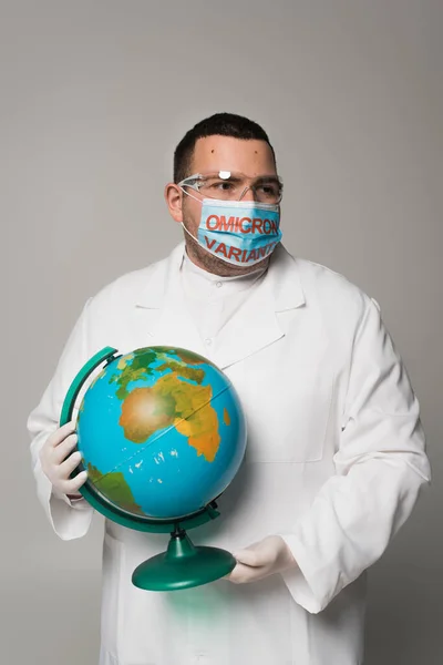 Scientist in medical mask with omicron variant lettering holding globe isolated on grey — Foto stock