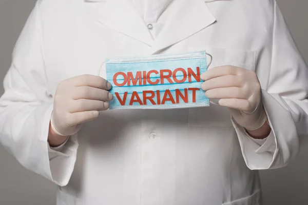 Cropped view of doctor in latex gloves holding medical mask with omicron variant lettering on grey background - foto de stock