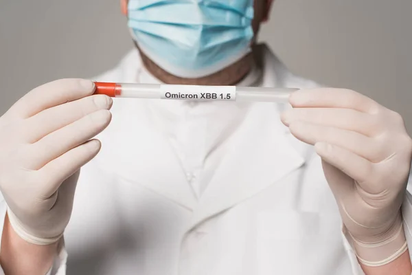Cropped view of blurred doctor in latex gloves holding throat swab with omicron xbb lettering isolated on grey — Foto stock