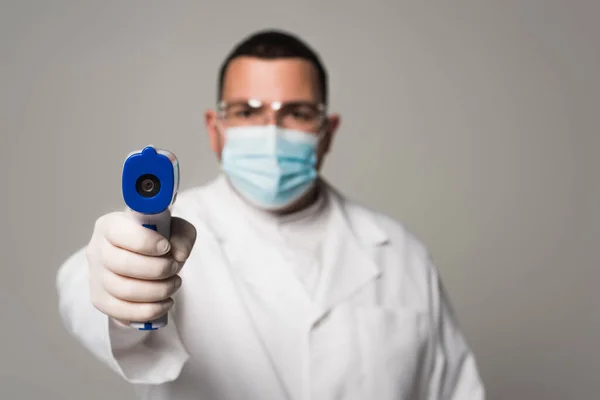 Médecin flou dans le masque médical tenant pyromètre isolé sur gris — Photo de stock