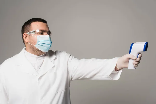 Doctor in protective goggles and mask holding pyrometer isolated on grey — Stock Photo