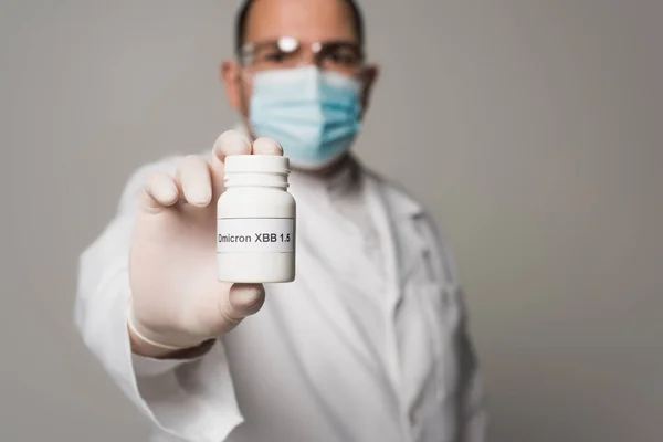 Blurred scientist in medical mask holding pills with omicron lettering isolated on grey - foto de stock