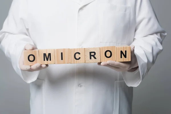 Cropped view of doctor in white coat and latex gloves holding wooden cubes with omicron lettering isolated on grey - foto de stock
