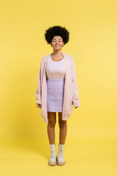 Full length of cheerful african american woman in cozy cardigan and skirt standing on yellow background — Stock Photo