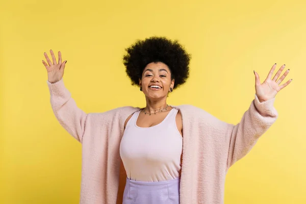 Optimistic african american woman with trendy hairstyle smiling at camera and waving hands isolated on yellow - foto de stock