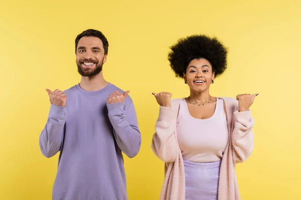 Cheerful interracial couple pointing with thumbs and looking at camera isolated on yellow — Stockfoto