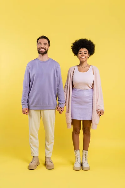 Full length of trendy interracial couple holding hands and smiling at camera on yellow background - foto de stock