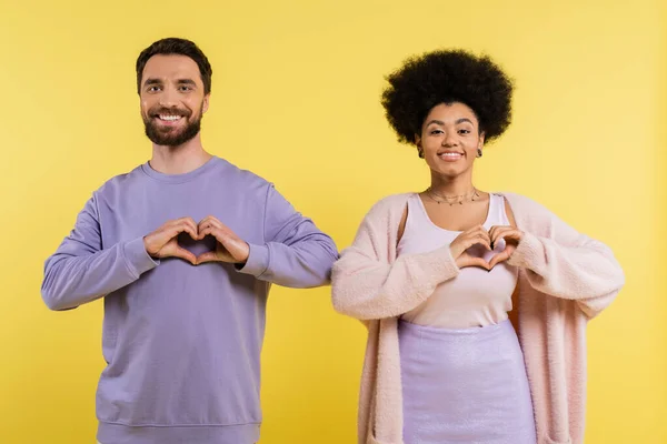 Счастливая многонациональная пара смотрит в камеру и показывает сердечные знаки изолированы на желтый — стоковое фото