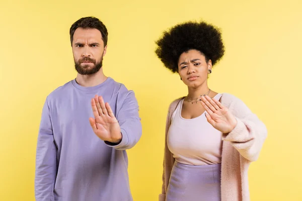 Displeased and frowning multiethnic couple looking at camera and showing refuse gesture isolated on yellow — Photo de stock