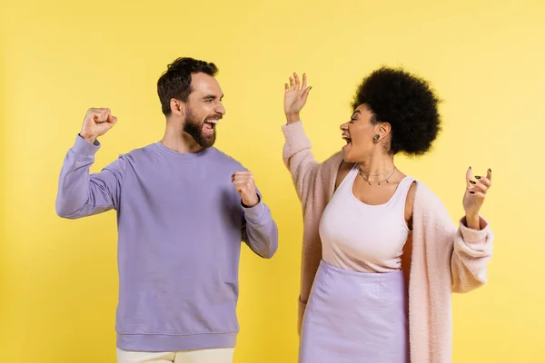 Overjoyed multiethnic couple screaming and showing win gesture while looking at each other isolated on yellow — Stockfoto