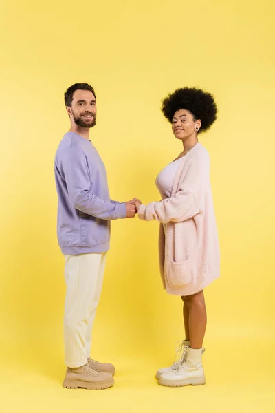 Полная длина межрасовой пары в стильной одежде, держась за руки и глядя на камеру на желтом фоне — стоковое фото
