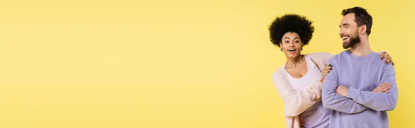 Cheerful african american woman hugging shoulders of smiling man standing with crossed arms isolated on yellow, banner — Stock Photo