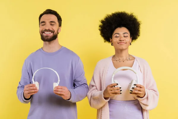 Pleased multiethnic couple with closed eyes holding wireless headphones isolated on yellow — Photo de stock