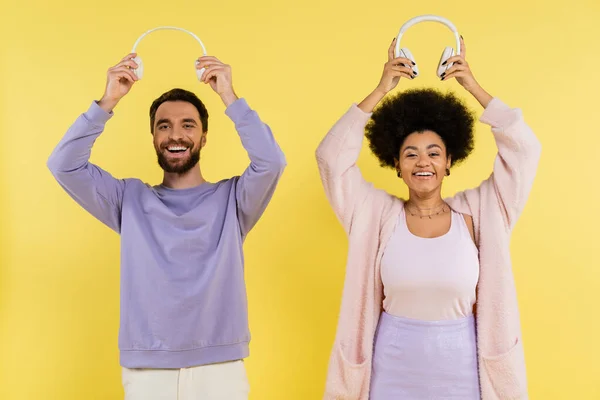 Cheerful and trendy interracial couple holding wireless headphones above heads isolated on yellow — Stock Photo