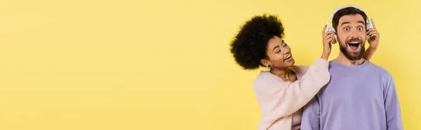 Curly african american woman putting wireless headphones on amazed man isolated on yellow, banner — Stock Photo