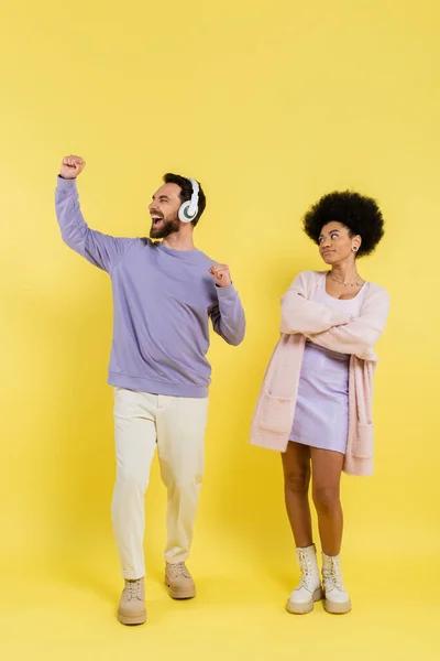 Full length of excited man in headphones rejoicing near skeptical african american woman standing with crossed arms on yellow — Stock Photo
