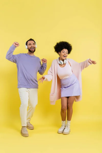 Full length of cheerful interracial couple in trendy clothes and wireless headphones dancing on yellow — Foto stock