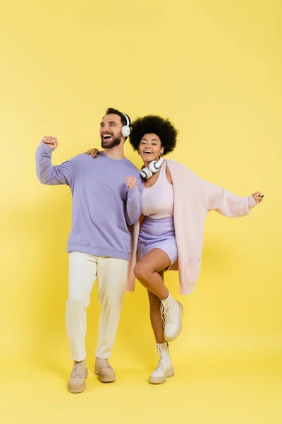 Full length of excited and stylish multiethnic couple dancing in wireless headphones on yellow background — Fotografia de Stock