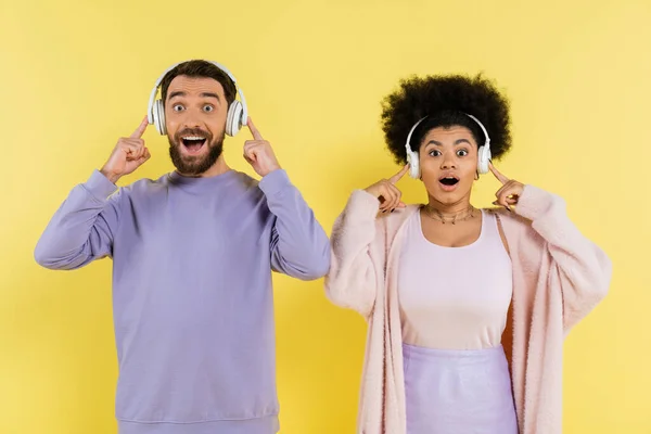 Amazed multiethnic couple listening music in wireless headphones and looking at camera isolated on yellow — Stock Photo
