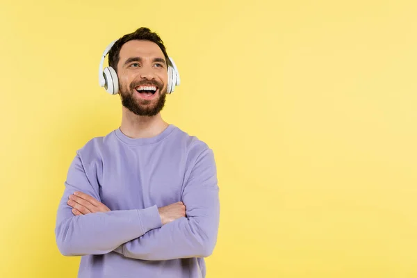 Hombre barbudo escuchando música en auriculares inalámbricos mientras está de pie con los brazos cruzados y riendo aislado en amarillo - foto de stock