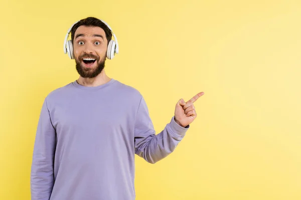 Amazed man in wireless headphones pointing with finger and looking at camera isolated on yellow — Foto stock