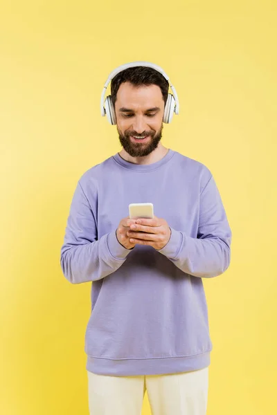 Smiling bearded man in purple pullover and wireless headphones using smartphone isolated on yellow — Stockfoto