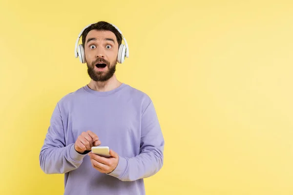 Erstaunter Mann mit drahtlosen Kopfhörern, Handy in der Hand, während er die Kamera isoliert auf gelb anschaut — Stockfoto