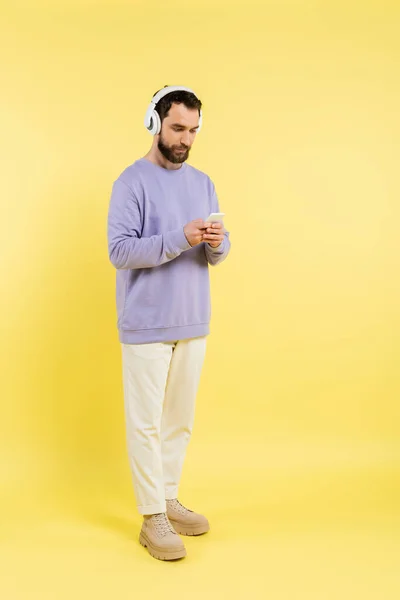 Full length of trendy bearded man in wireless headphones using mobile phone on yellow — Stock Photo