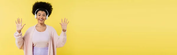 Радостная африканская американка слушает музыку в беспроводных наушниках и машет руками изолированными на желтом, баннер — стоковое фото
