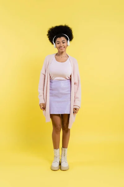 Full length of cheerful african american woman in cardigan and wireless headphones looking at camera on yellow background — Stockfoto