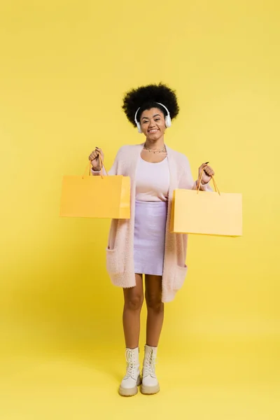 Full length of happy african american woman in wireless headphones and boots holding shopping bags on yellow background — Foto stock