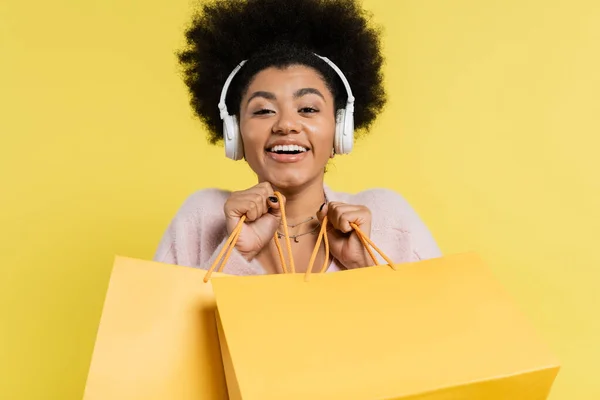 Excited african american woman in wireless headphones holding shopping bags and looking at camera isolated on yellow — Stockfoto