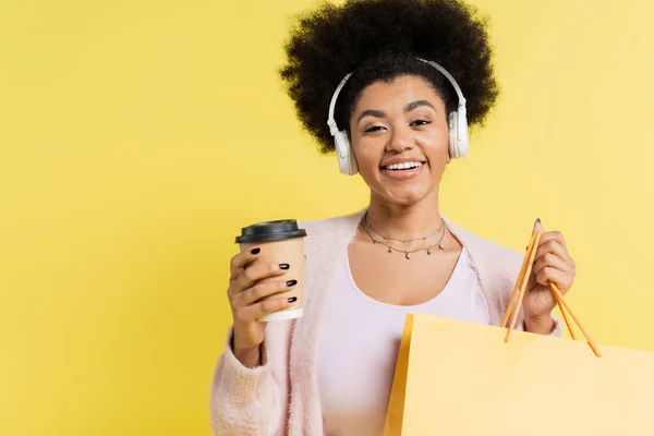 Positive african american woman in wireless headphones standing with coffee to go and shopping bag isolated on yellow - foto de stock
