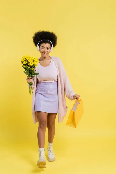 Full length of smiling african american woman in wireless headphones walking with flowers and shopping bag on yellow background — Fotografia de Stock