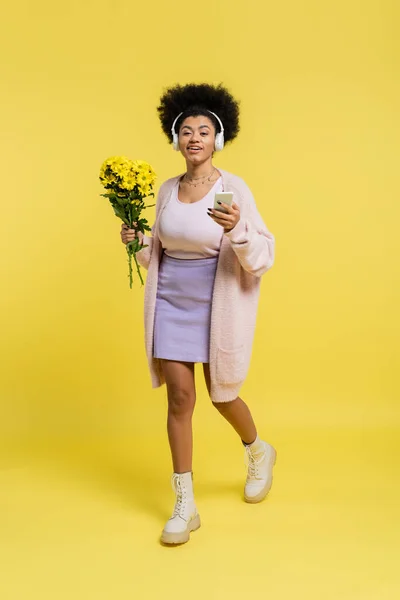 Full length of happy african american woman in wireless headphones walking with bouquet and smartphone on yellow — Fotografia de Stock