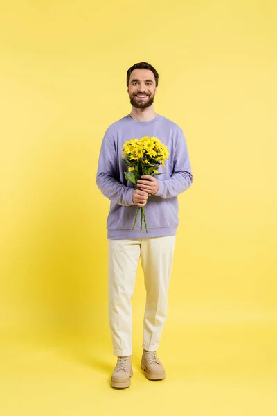 Full length of bearded and trendy man holding bouquet and smiling at camera on yellow - foto de stock