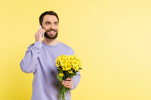 Smiling man in purple pullover holding flowers and talking on smartphone isolated on yellow — Stockfoto