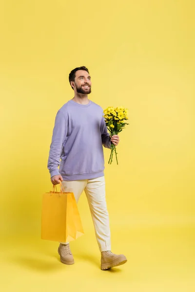 Pleine longueur de l'homme heureux et élégant marche avec des sacs à provisions et des fleurs sur fond jaune — Photo de stock