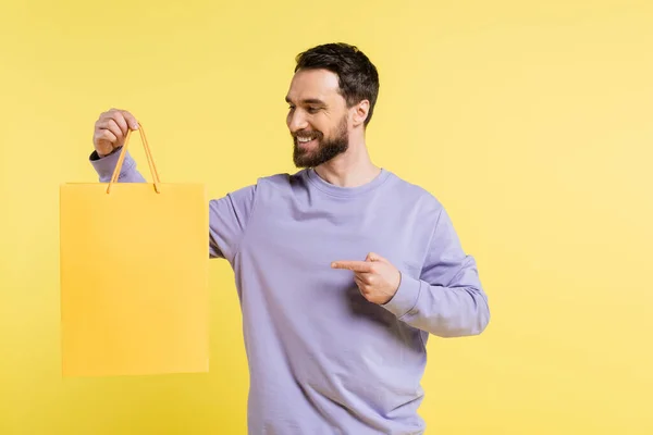 Joyful man in purple pullover pointing with finger at shopping bag isolated on yellow — Stockfoto
