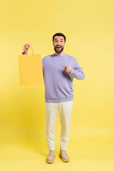 In voller Länge auf aufgeregtem und trendigem Mann, der in die Kamera blickt und auf Einkaufstasche auf gelbem Hintergrund zeigt — Stockfoto