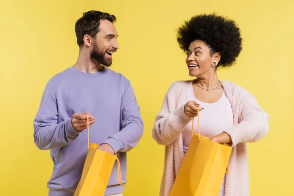 Bearded man and curly african american woman holding shopping bags and smiling at each other isolated on yellow — Fotografia de Stock