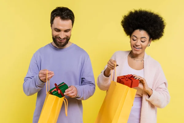 Happy interracial couple in stylish clothes holding shopping bags and presents isolated on yellow — Stockfoto