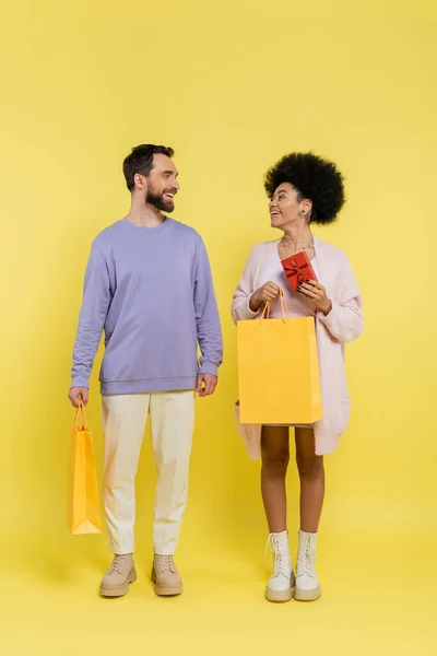 Full length of trendy african american woman holding gift box and shopping bag near smiling bearded man on yellow — Foto stock