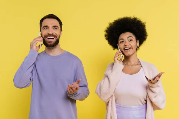 Trendy and positive interracial couple gesturing while talking on mobile phones isolated on yellow — Stockfoto
