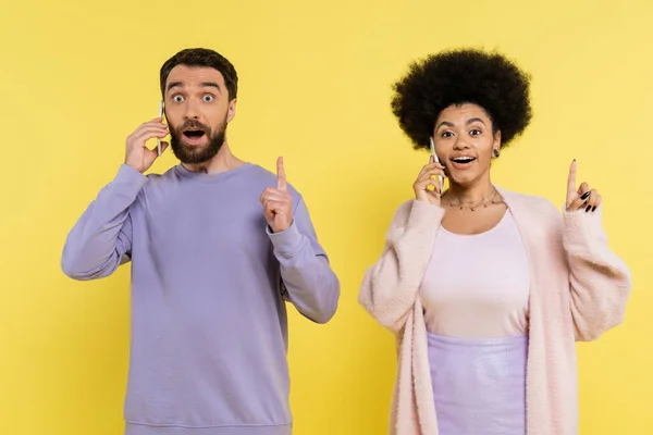 Amazed multiethnic couple showing idea gesture while talking on smartphones isolated on yellow — Stock Photo